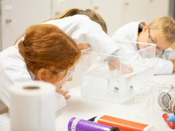 Drei Kinder in Laborkitteln beobachten und experimentieren mit Objekten, die in einem transparenten Wassertank auf einem Labortisch in Wasser versenkt sind.
