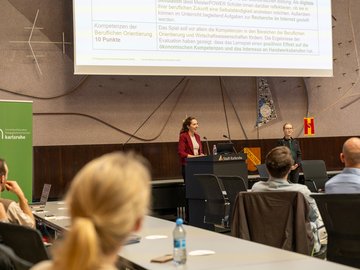Vortrag von Marie Tuchscherer (l.) und Prof. Claudia Wiepcke. Am Rednerpult mit der Leinwand über sich Frau Tuchscherer. Im vordergrund Publikum.er.