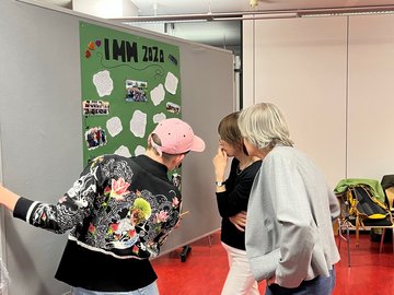 Drei Personen betrachten ein Poster mit dem Titel IMM 2020.