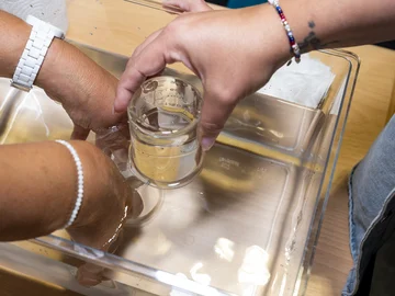 Eine Hand stellt ein Becherglas in ein Gefäß, zwei andere Hände helfen.