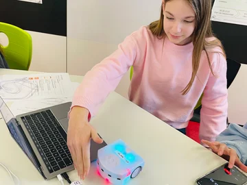 Eine Schülerin arbeitet mit einem kleinen Roboter, der auf einem Tisch steht; daneben stehen ein Laptop, eine Ladestation und ein Smartphone.