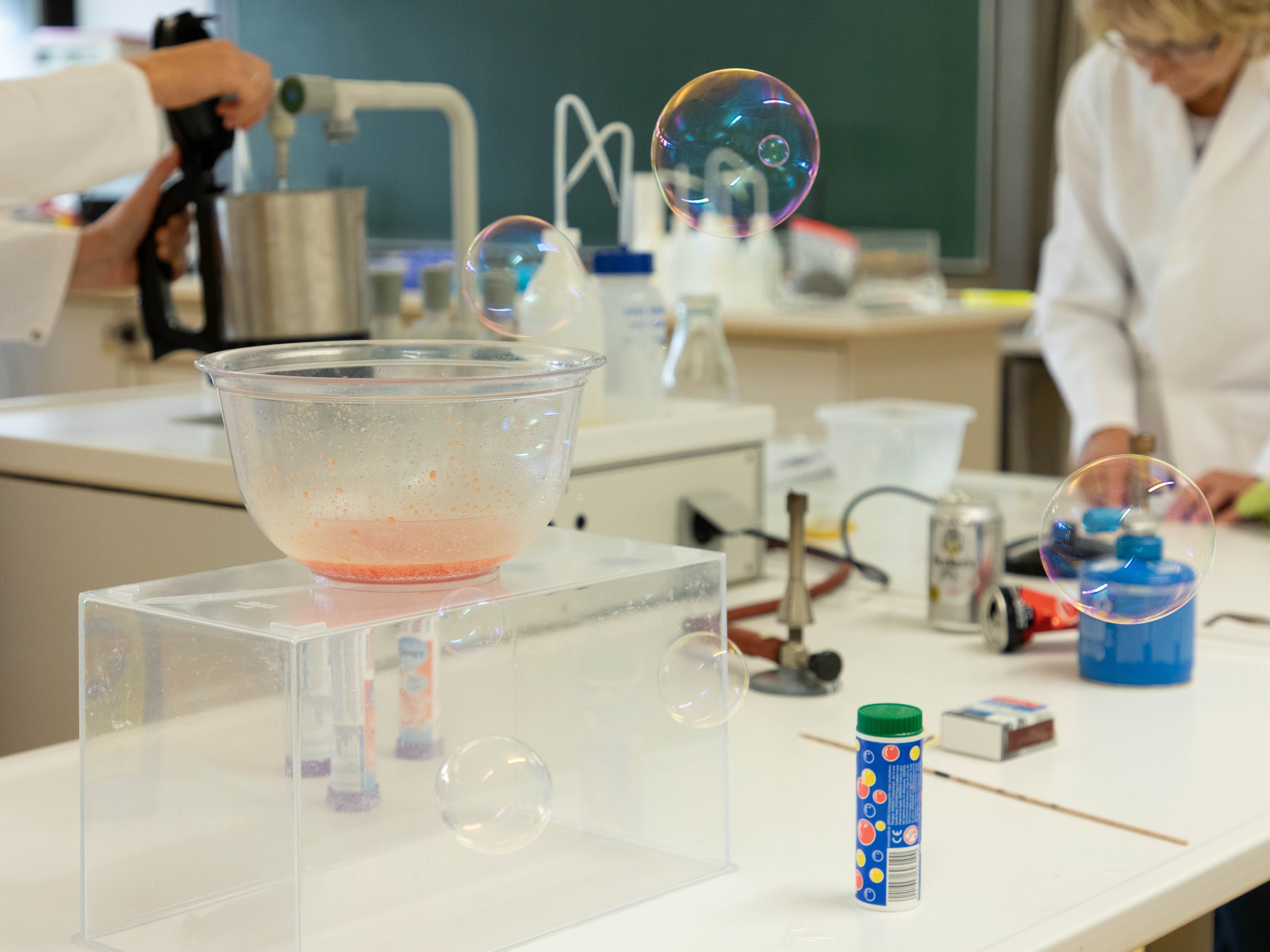 Ein chemischer Versuchsaufbau im Labor. In der Luft schweben Seifenblasen. Im Hintergrund arbeiten Menschen in Laborkitteln.