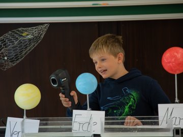 Ein Junge steht hinter Planetenmodellen von Venus, Erde und Mars und hält ein technisches Gerät.