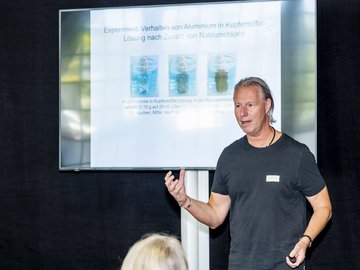 Ein Vortragender deutet auf den Bildschirm und erklärt die aktuelle Folie zum Experiment Verhalten von Aluminium in Kupfersulfatlösung nach Zusatz von Natriumchlorid.