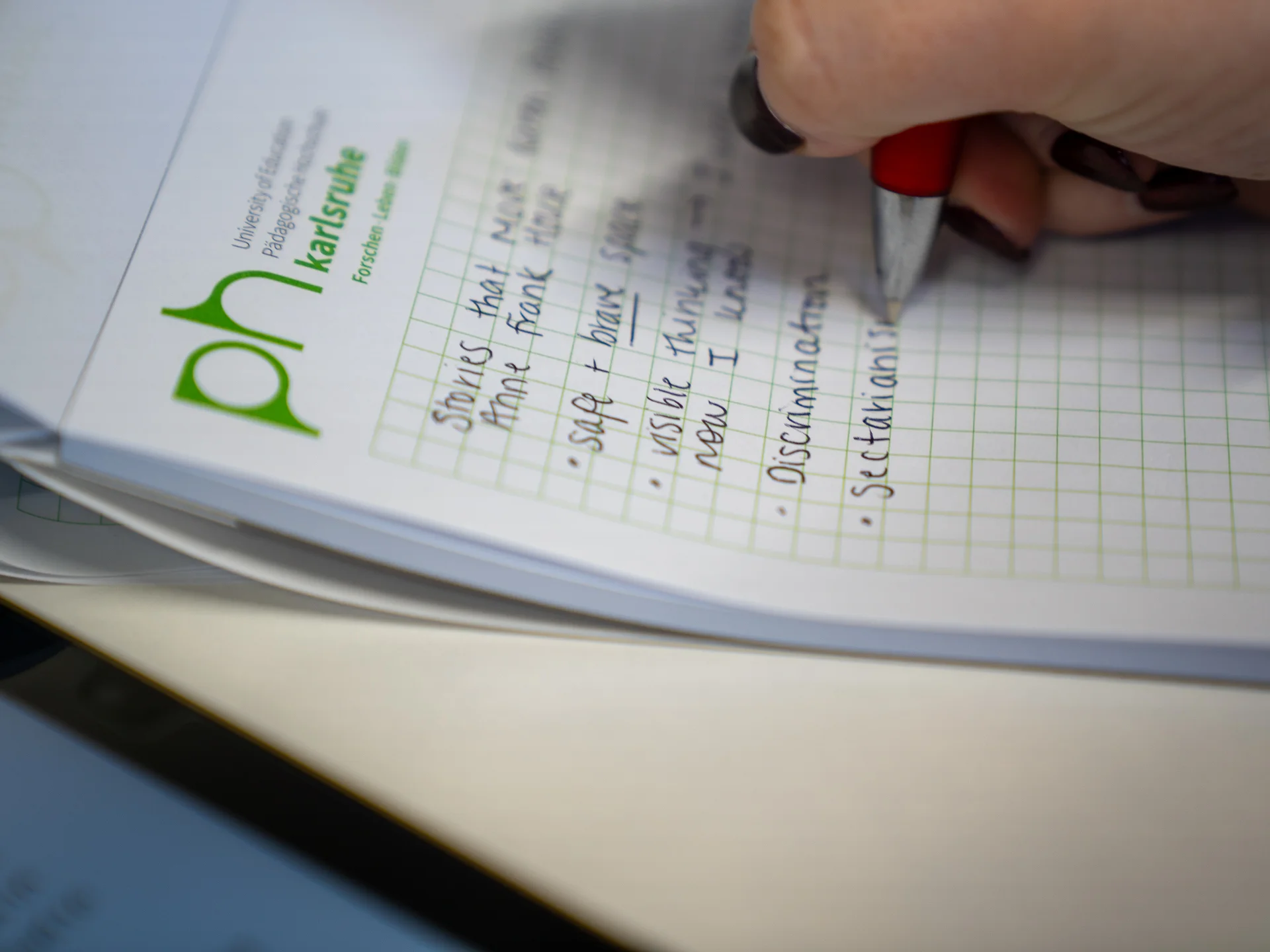 A hand writing on a notebook that has the logo of Karlsruhe University of Education on it.