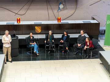 Diskussionsrunde mit PHKA-Wissenschaftler:innen auf Stühlen vor dem Pult im Bürgersaal der Stadt Karlsruhe. dei Moderatorin steht.