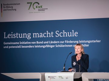 Die Vortragende spricht auf dem Podium, dahinter ein Banner mit Logos und Schriftzug Leistung macht Schule.