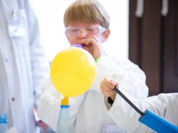 Ein Junge in Laborkittel und Schutzbrille bläst einen blauen Ballon auf. Vor ihm steht ein luftgefüllter gelber Ballon, der an einer Falsche befestigt ist.