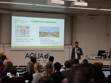 Marc Philipp Janson bei der Eröffnung der AQUA-d Konferenz 2025. Er steht neben der Leinwand; im Vordergrund sind Zuhörende in den Sitzeihen von hinten zu sehen. 