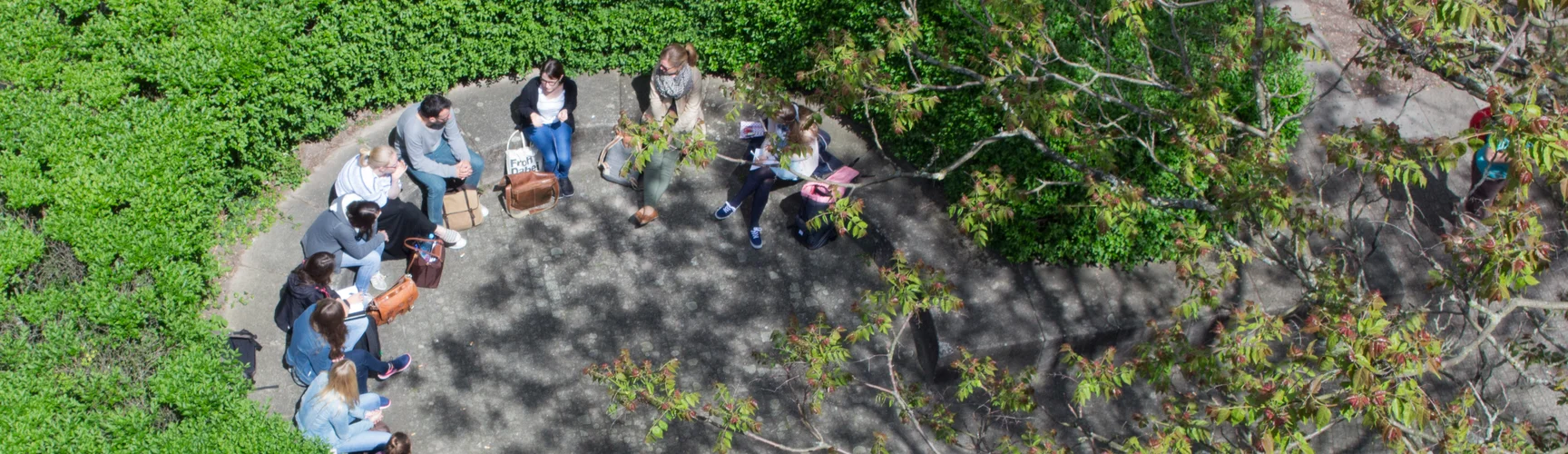 Eine Gruppe Personen sitzt auf dem Campus in einem durch eine Hecke geschützten Bereich