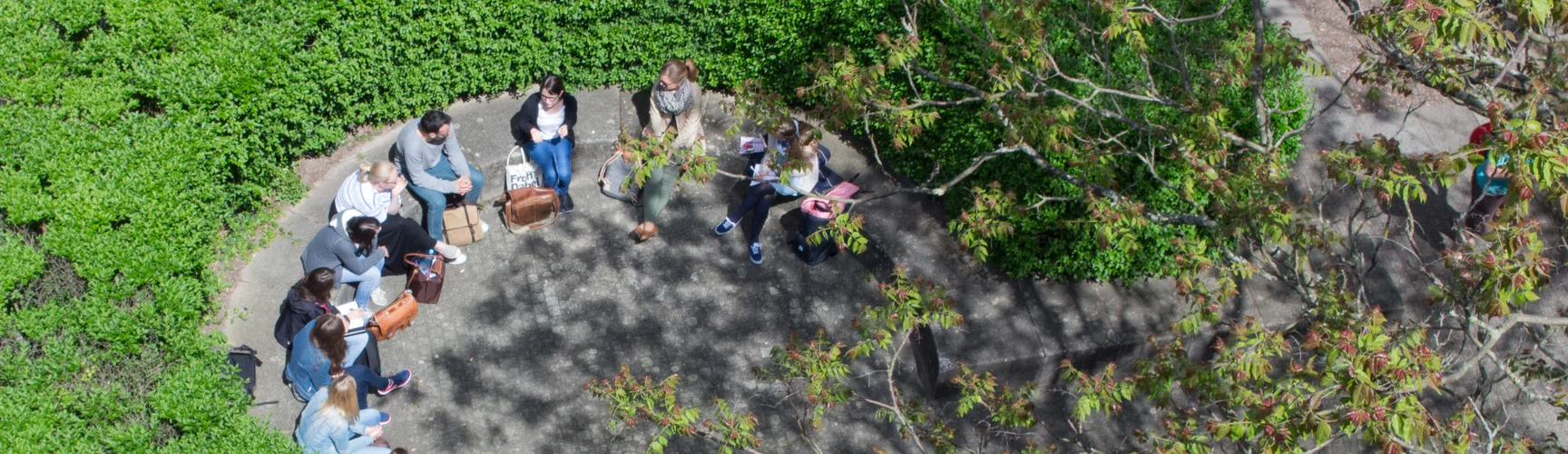 Eine Gruppe Personen sitzt auf dem Campus in einem durch eine Hecke geschützten Bereich