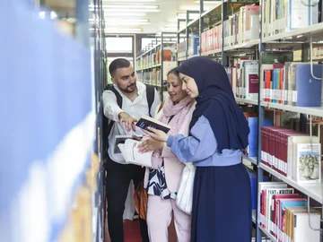 Ein Mann und zwei Frauen, eine davon trägt ein Kopftuch, stehen in der Bibliothek und betrachten ein Buch.