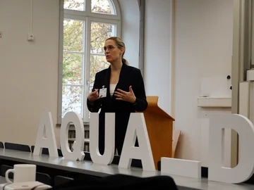 Anique de Bruin steht in der Mitte des Bildes und hält eine Keynote. Im Vordergrund sind weiße Holzbuchstaben (AQUA-D) auf einem Tisch in einem Hörsaal.