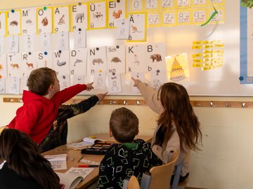 Vier Kinder im Klassenzimmer zeigen mit ihren Fingern auf eine Pinnwand mit Bildern von Tieren und deren Anfangsbuchstaben