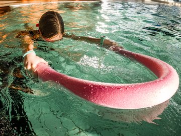 Ein Kind schwimmt in einem Schwimmbecken, es hat das Gesicht unter Wasser und hält eine Schwimmnudel vor sich.