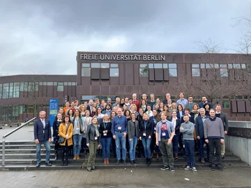 Eine Gruppe von Personen steht vor einem Gebäude der Freien Universität Berlin auf einer Treppe, einige tragen Namensschilder.