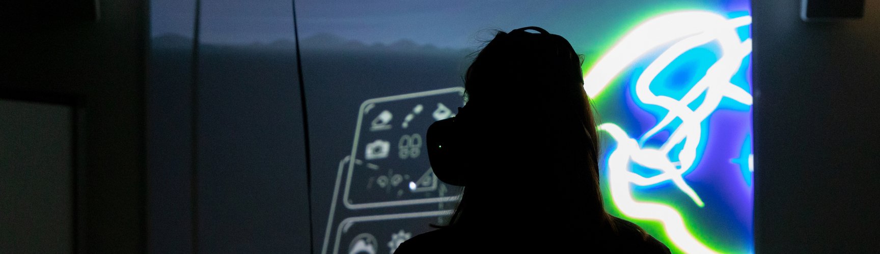 Eine Person mit einer VR Brille steht in einem dunklen Raum und schaut auf einen erleuchteten Monitor