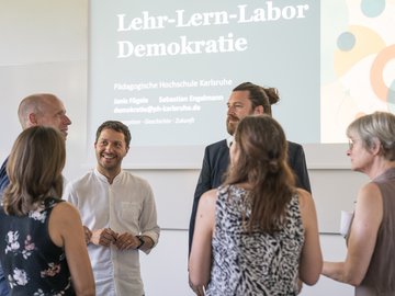 Vor einer Leinwand mit gezeigter Folie unterhalten sich angeregt drei Männer und drei Frauen.