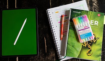 Symbolbild KI in der Schule Ein Tablet und ein Schulbuch liegen symbolisch nebeneinander. Das Schubuch liegt zudem auf einem Block und es gibt einige bunte Stiftte.