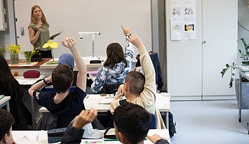 Unterrichtsszene Eine Lehrerin steht vor einer Schulklasse. Dei Schülerinnen und Schüler melden sich teilweise und sind von hinten zu sehen.