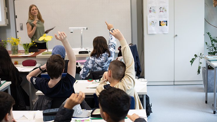 Eine Lehrerin steht vor einer Schulklasse. Dei Schülerinnen und Schüler melden sich teilweise und sind von hinten zu sehen.