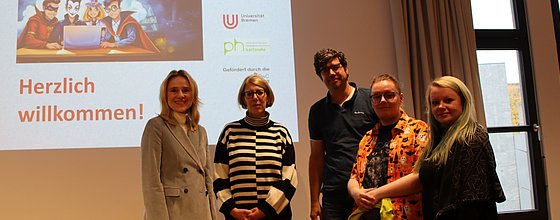 Dei Projketpartner, vier Frauen und ein Mann, stehen vor dem Titelbild der PPT für den Kick-Off des Projekts in einem Seminarraum. 