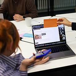 Schülerinnen vor einem Laptop und analogen Bildchen in der Hand beschäftigen sich mit KI im Kustunterrichtsich mit