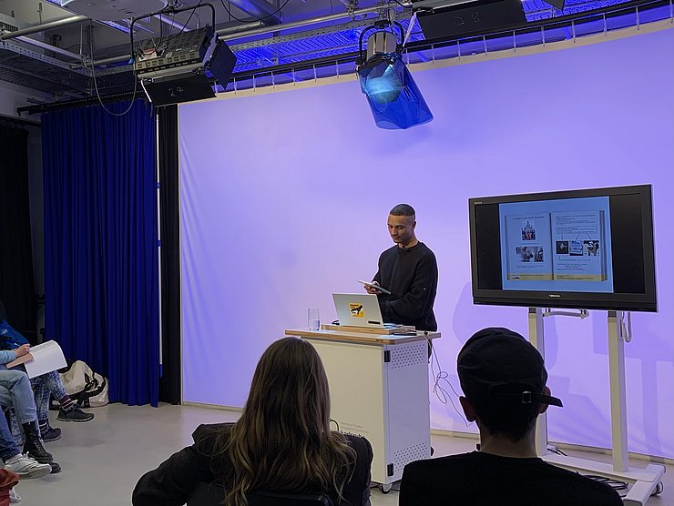 Eine Person hält einen Vortrag an einem Rednerpult in einem Studio, während mehrere Zuhörende sitzen und auf einen Bildschirm mit Präsentationsfolien schauen.