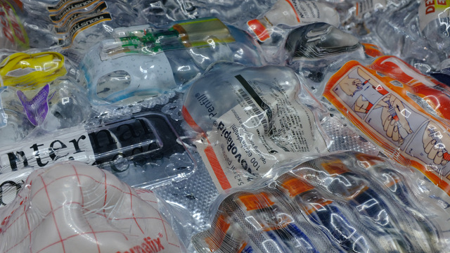 Das Bild zeigt eine Sammlung von medizinischen Verpackungen und Einwegmaterialien, die unter einer klaren, möglicherweise kunststoffartigen oder harzartigen Oberfläche eingeschlossen sind. Zu sehen sind beispielsweise:      Infusionsbeutel      Blisterverpackungen      Einweghandschuh-Verpackungen mit Anleitungen      Teile von Spritzen oder medizinischen Geräten      Verpackungsbeschriftungen in mehreren Sprachen  Das Arrangement wirkt bewusst gestaltet, fast skulptural – möglicherweise handelt es sich um ein Kunstwerk oder eine kritische Auseinandersetzung mit dem Thema medizinischer Müll, Sterilität und Wegwerfgesellschaft.